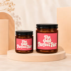 Two amber glass candles with pink labels that say "The Odd Perfect Pair Vowed".