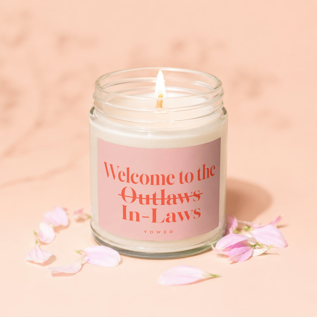Candle with lit wick reads "Welcome to the Outlaws In-Laws" on pink label.
