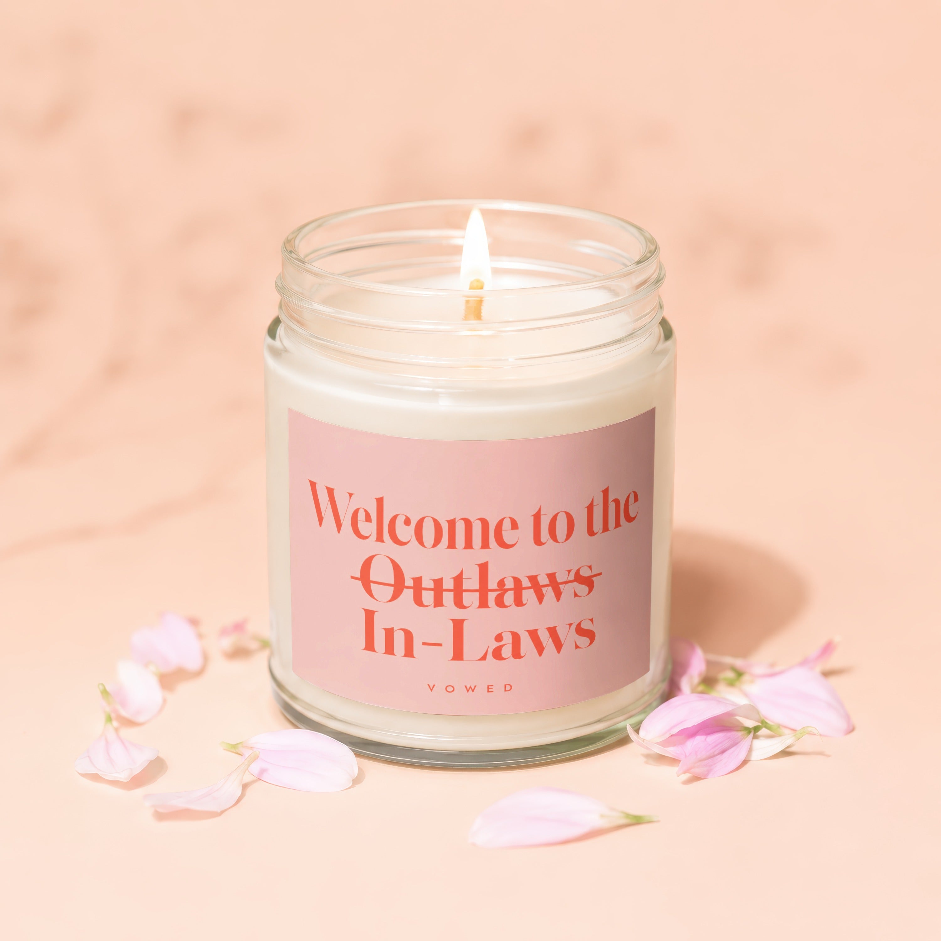 Candle with lit wick reads "Welcome to the Outlaws In-Laws" on pink label.