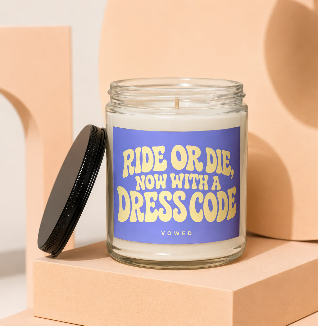 Clear glass candle with purple label reading "RIDE OR DIE, NOW WITH A DRESS CODE VOWED".
