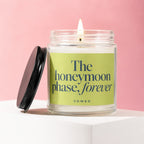 Glass jar candle with a lit wick on a white surface, reads "The honeymoon phase, forever".