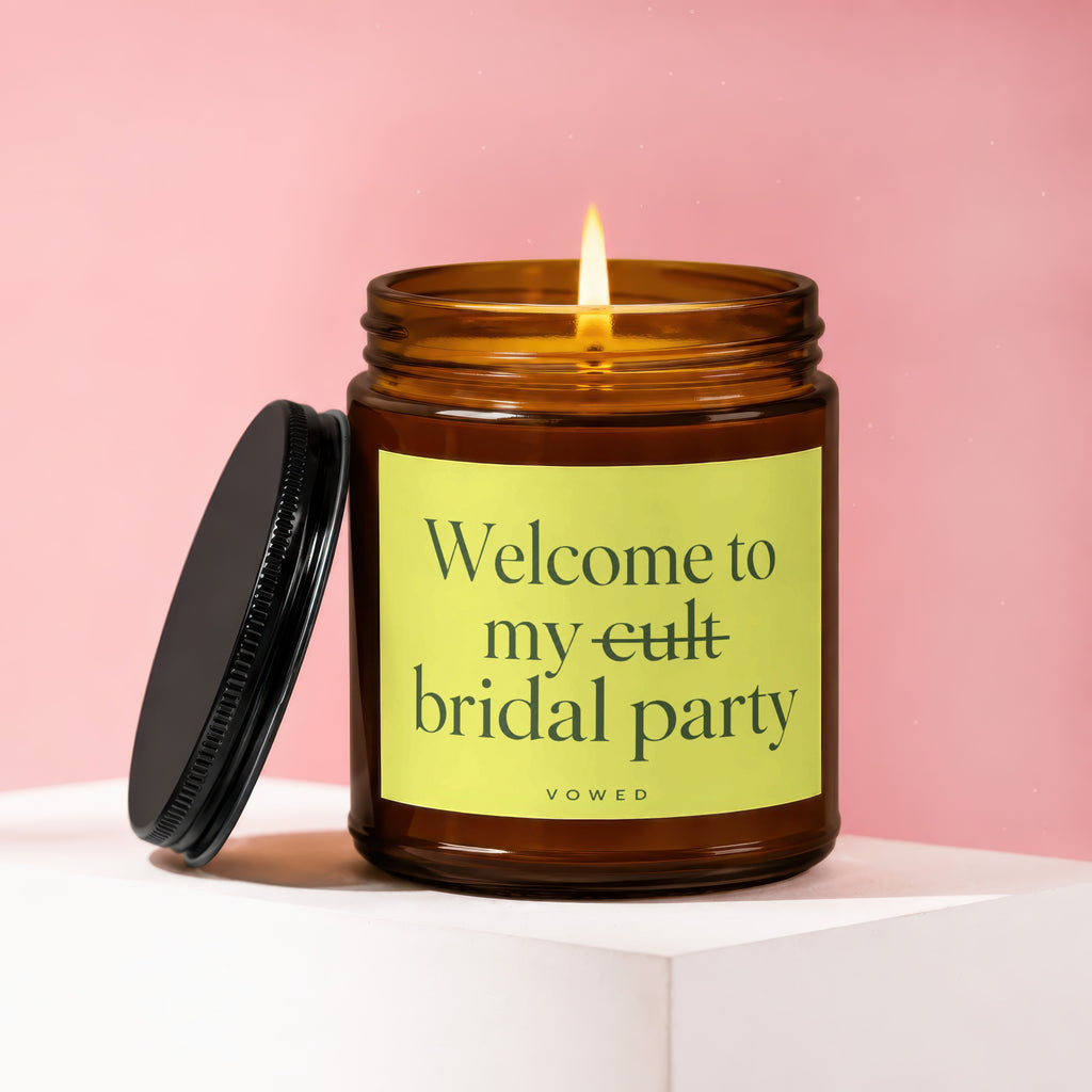 Amber glass candle with a lit wick and a black lid. Label reads "Welcome to my cult bridal party VOWED".