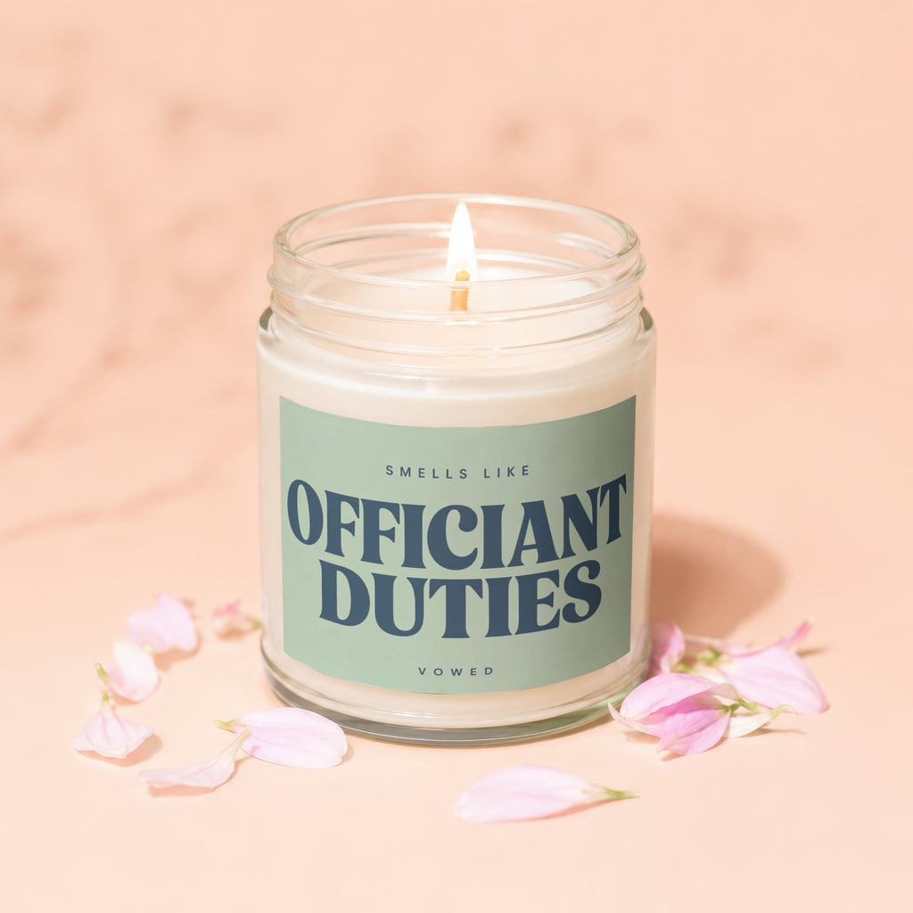 Glass jar candle labeled "Smells Like Officiant Duties" with a lit wick and petals.
