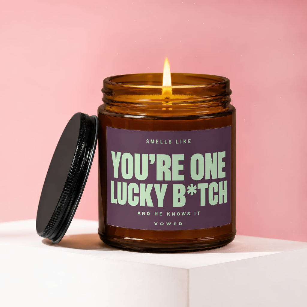 A lit candle in a brown glass jar with a purple label that reads "YOU'RE ONE LUCKY B*TCH".