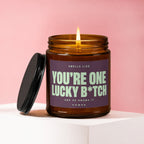 A lit candle in a brown glass jar with a purple label that reads "YOU'RE ONE LUCKY B*TCH".