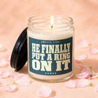 Glass candle with a lit wick, captioned "Smells Like He Finally Put a Ring On It Vowed."