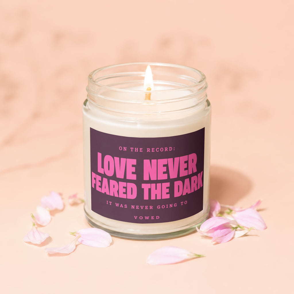 Candle with pink text "Love Never Feared The Dark" on label, surrounded by flower petals.