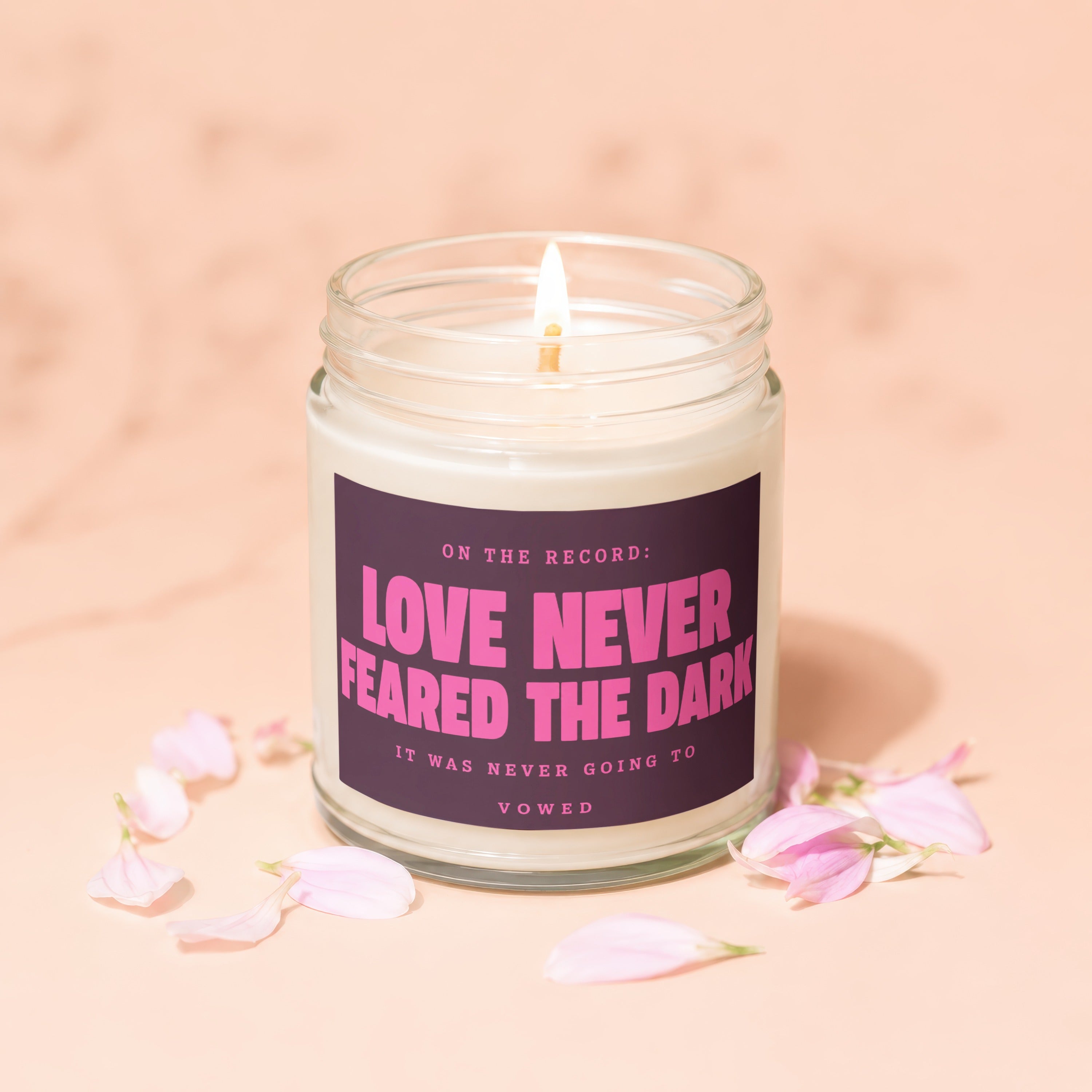 Candle with pink text "Love Never Feared The Dark" on label, surrounded by flower petals.