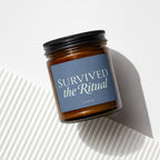 Amber glass candle with "Survived the Ritual" and "VOWED" text on a blue label.