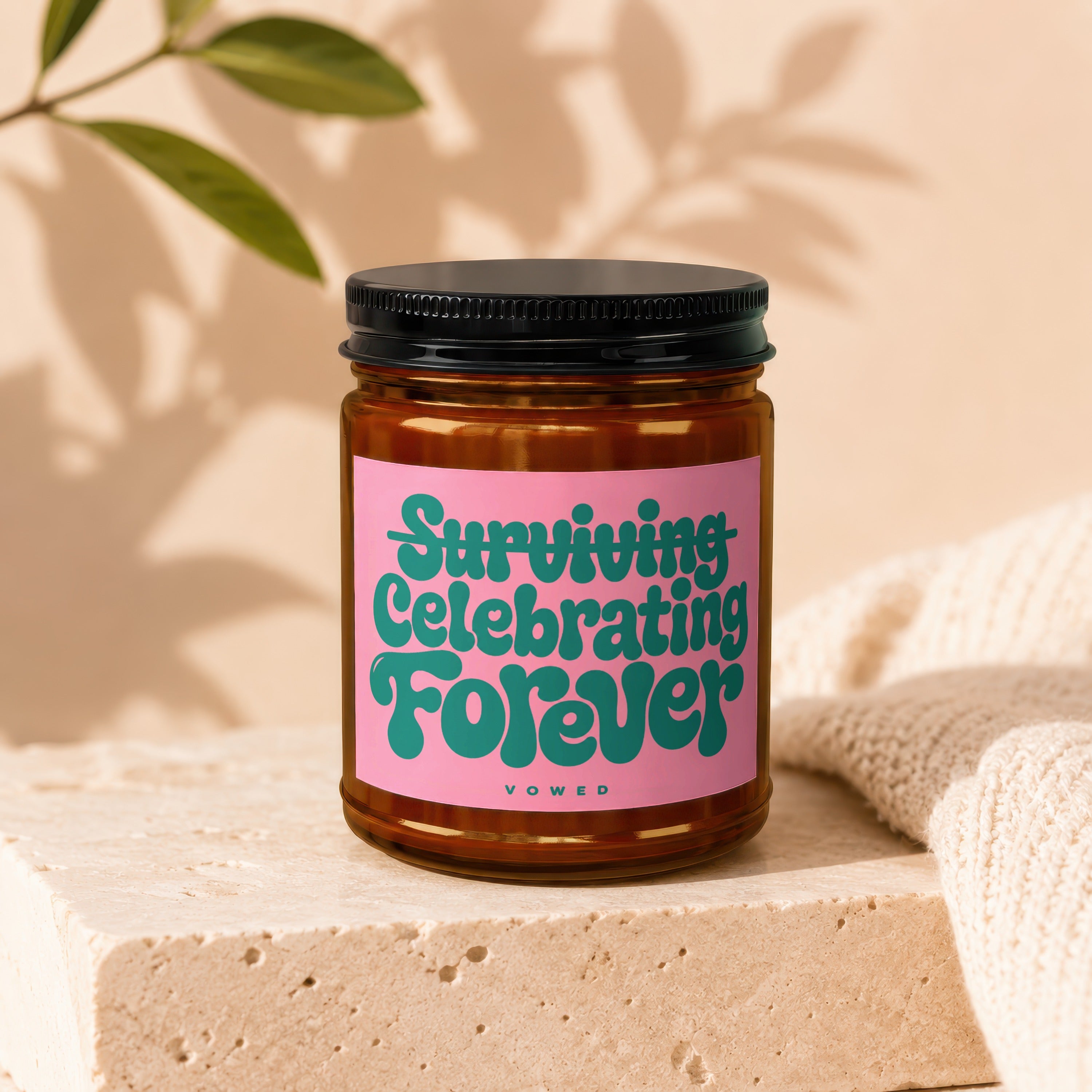 Amber glass candle on stone block with pink label text "Surviving Celebrating Forever".