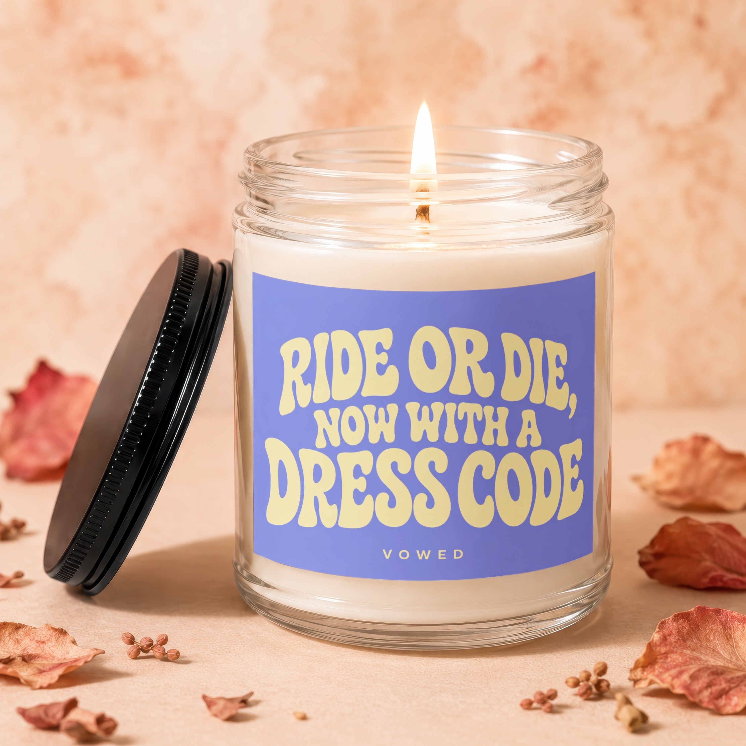 Glass candle with lid nearby, lit wick, and purple label reading "Ride Or Die, Now With A Dress Code".