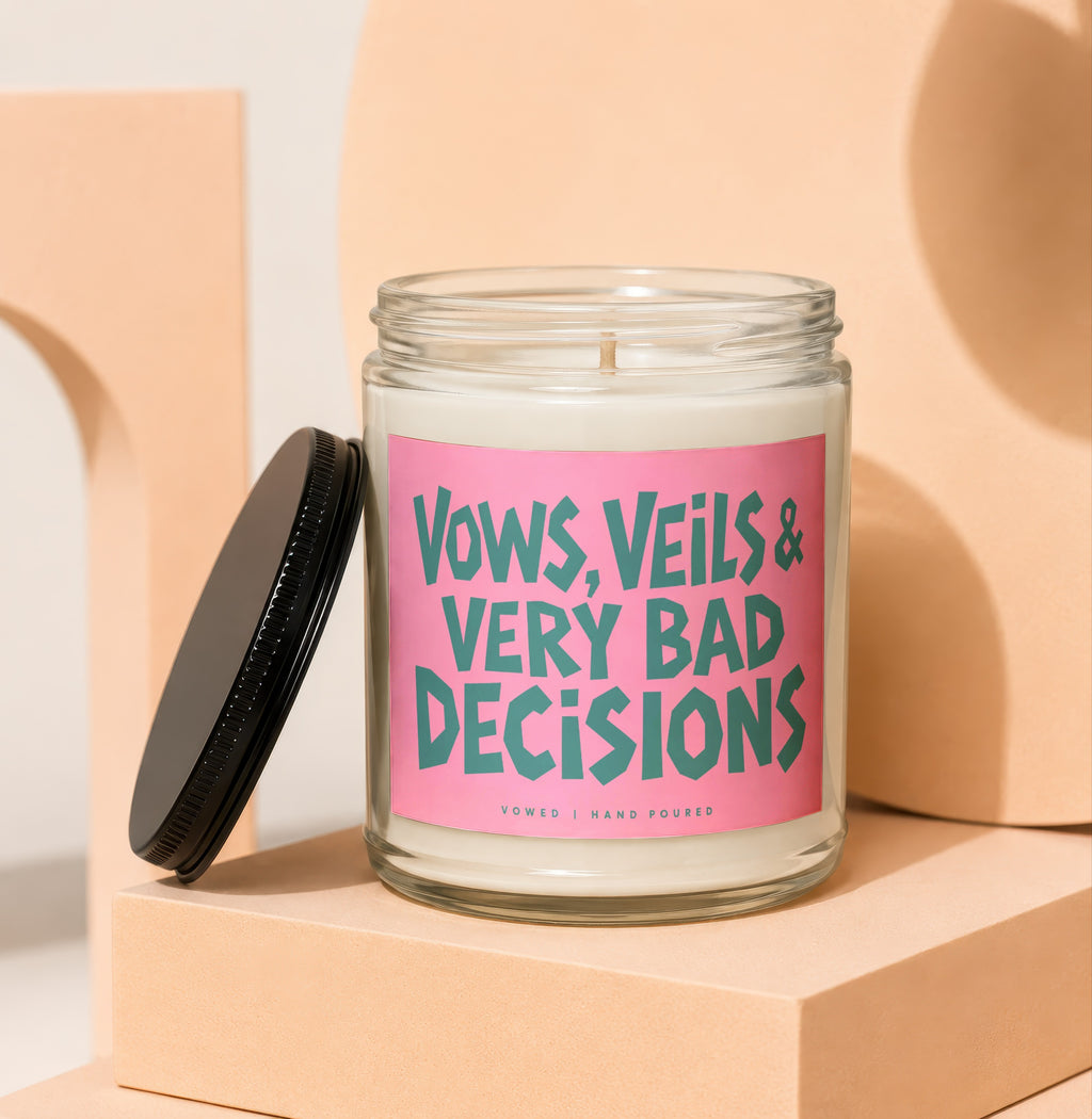Glass jar candle with pink label reading "Vows, Veils & Very Bad Decisions."