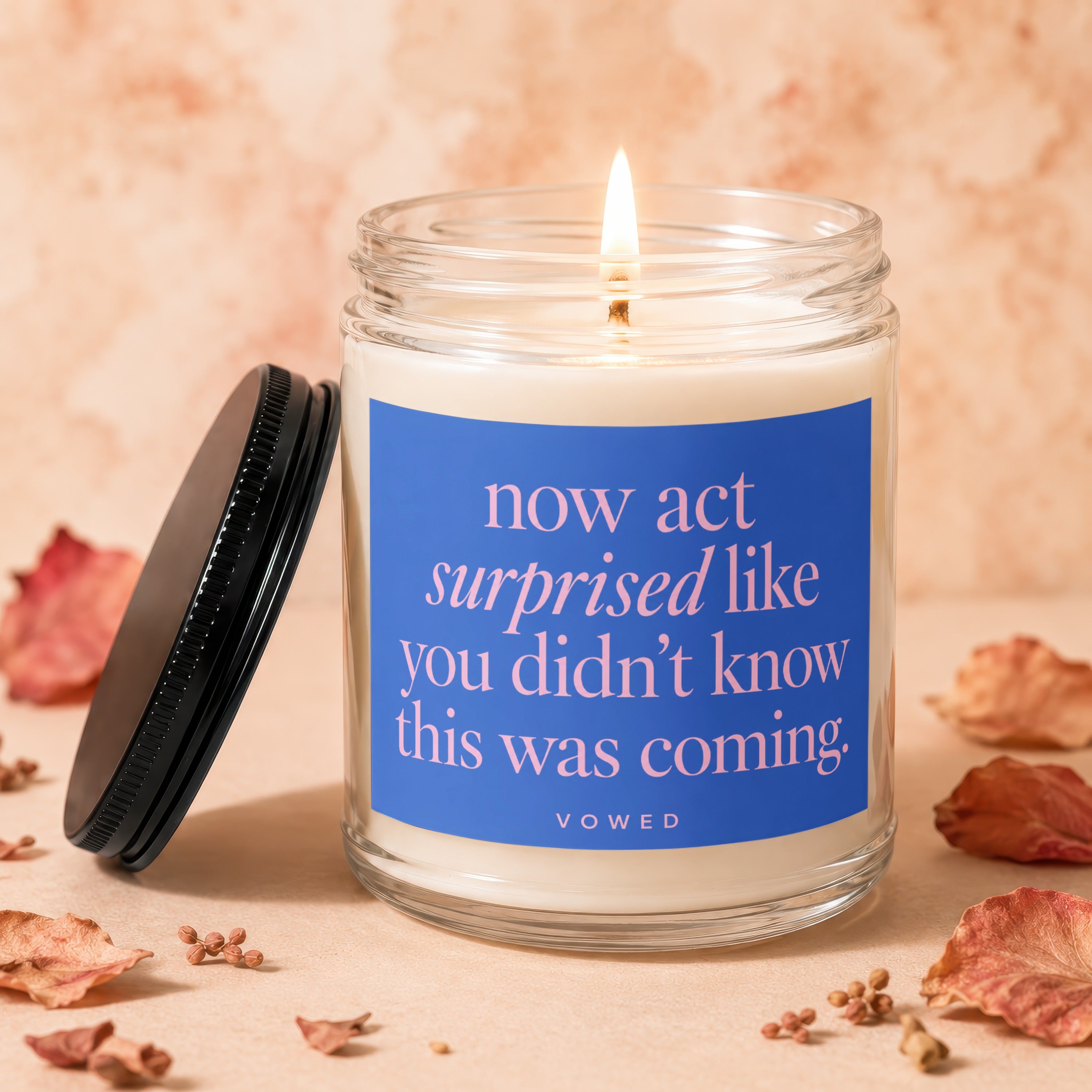 Lit candle in glass jar with blue label reading "now act surprised like you didn't know this was coming."