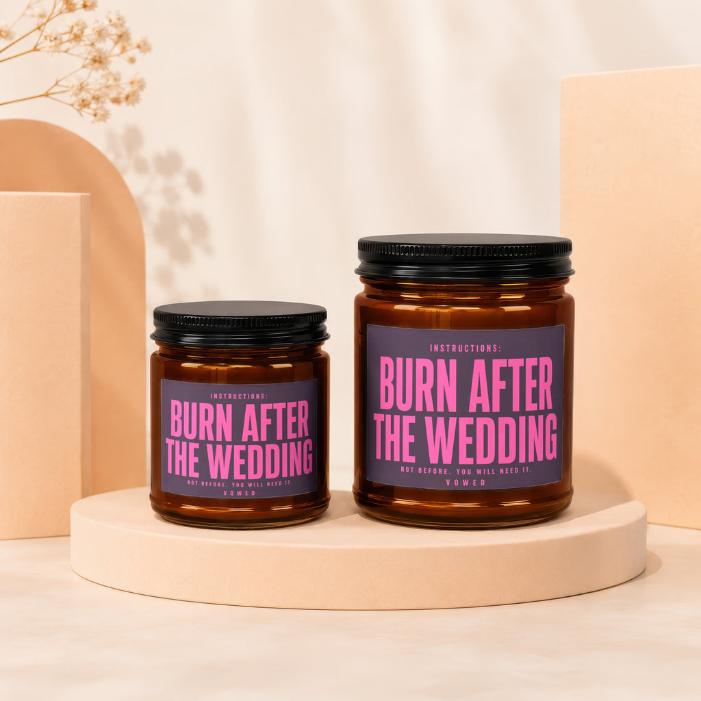 Two amber glass candles with black lids and pink text reading "Burn After The Wedding".