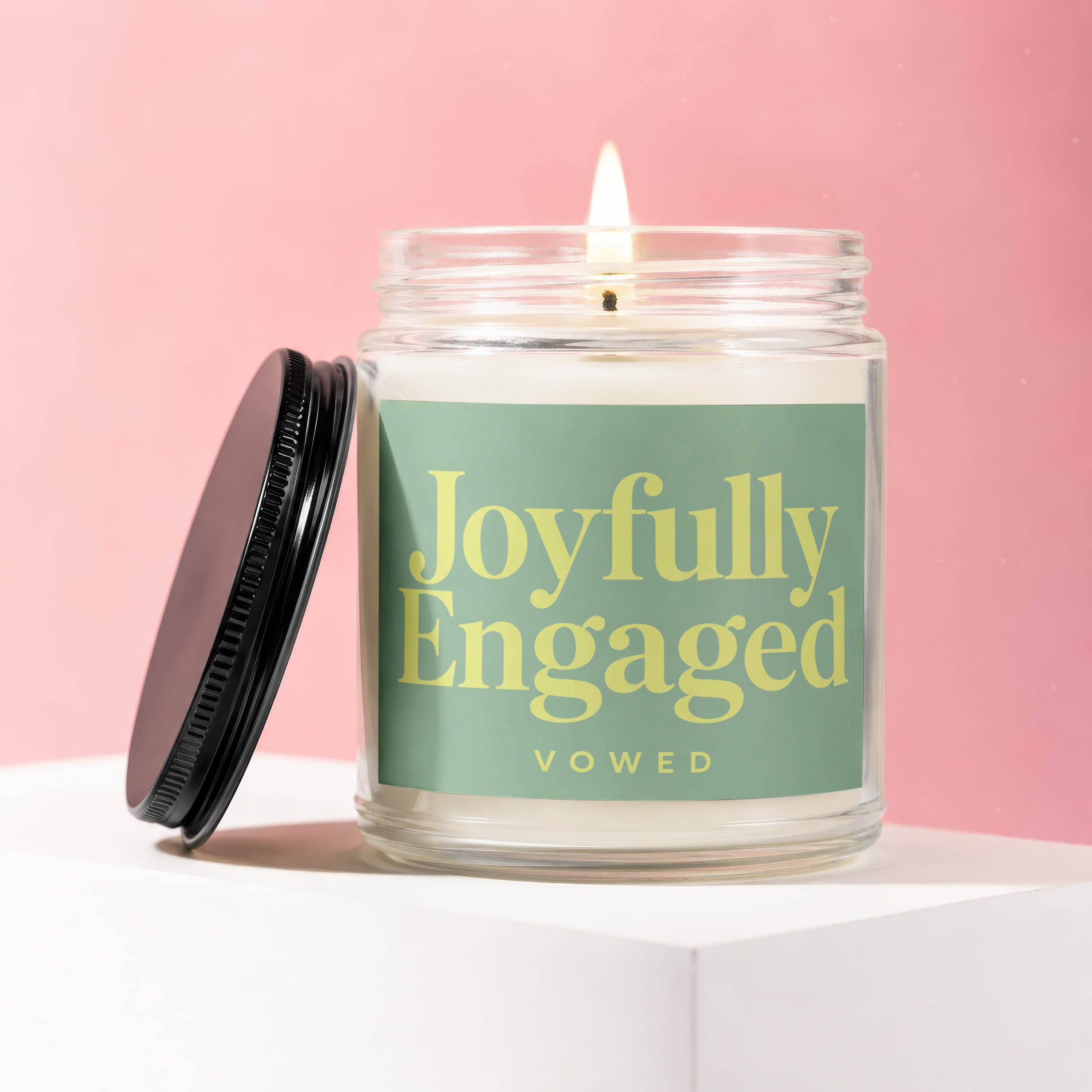Lit glass jar candle with green label reading "Joyfully Engaged" on a white surface.