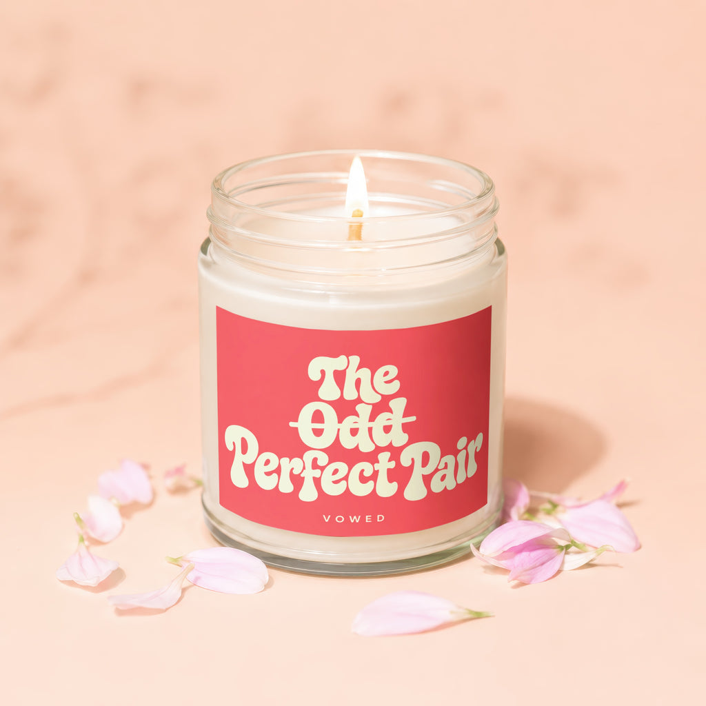 Lit candle in a glass jar with a pink label reading "The Odd Perfect Pair" below "VOWED".