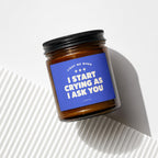 Amber glass candle with blue label reading "Light Me When I Start Crying As I Ask You".
