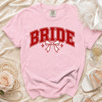 Pink t-shirt with "BRIDE" in red collegiate font and a white bow.