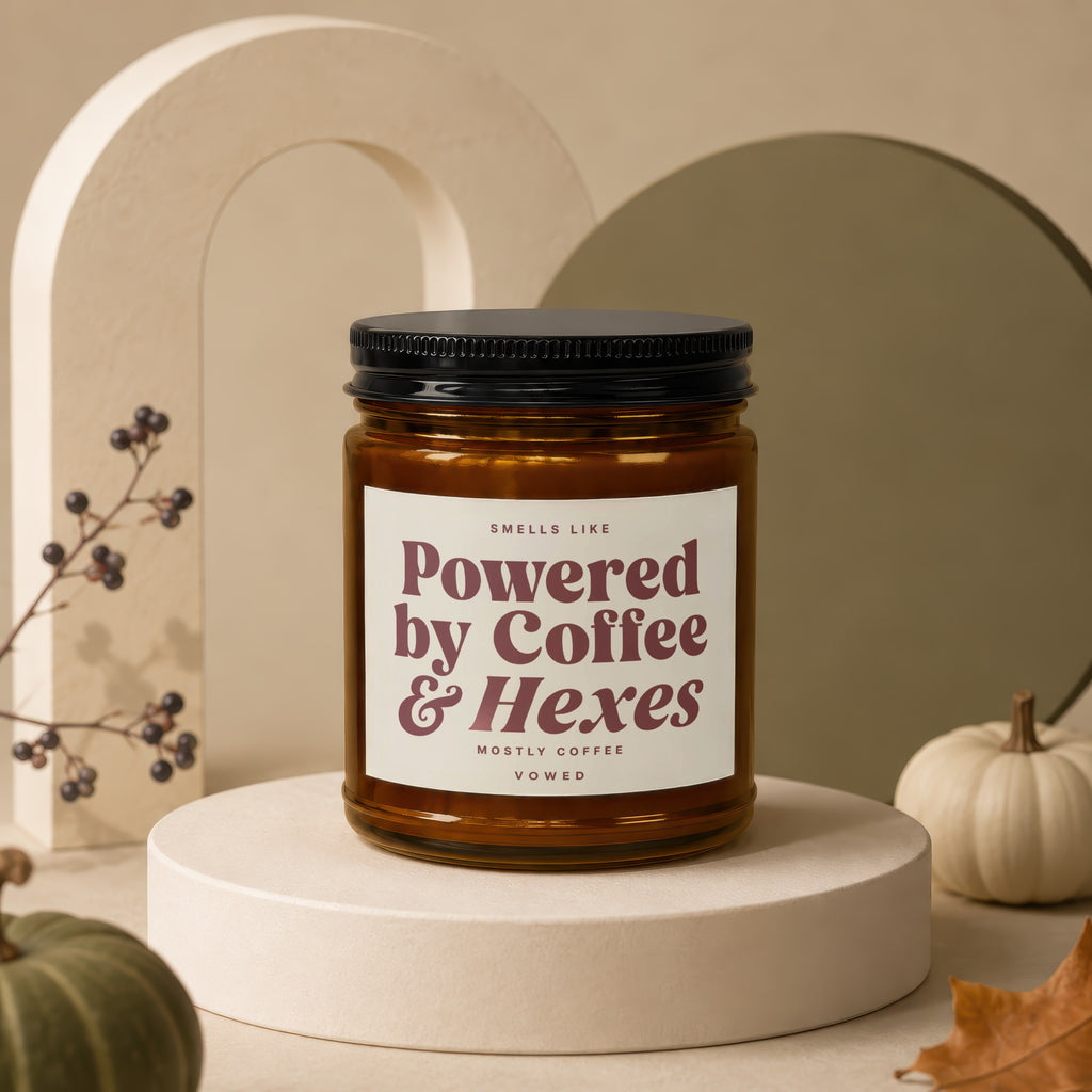 Brown glass candle labeled "Powered by Coffee & Hexes" on a beige pedestal with pumpkins.