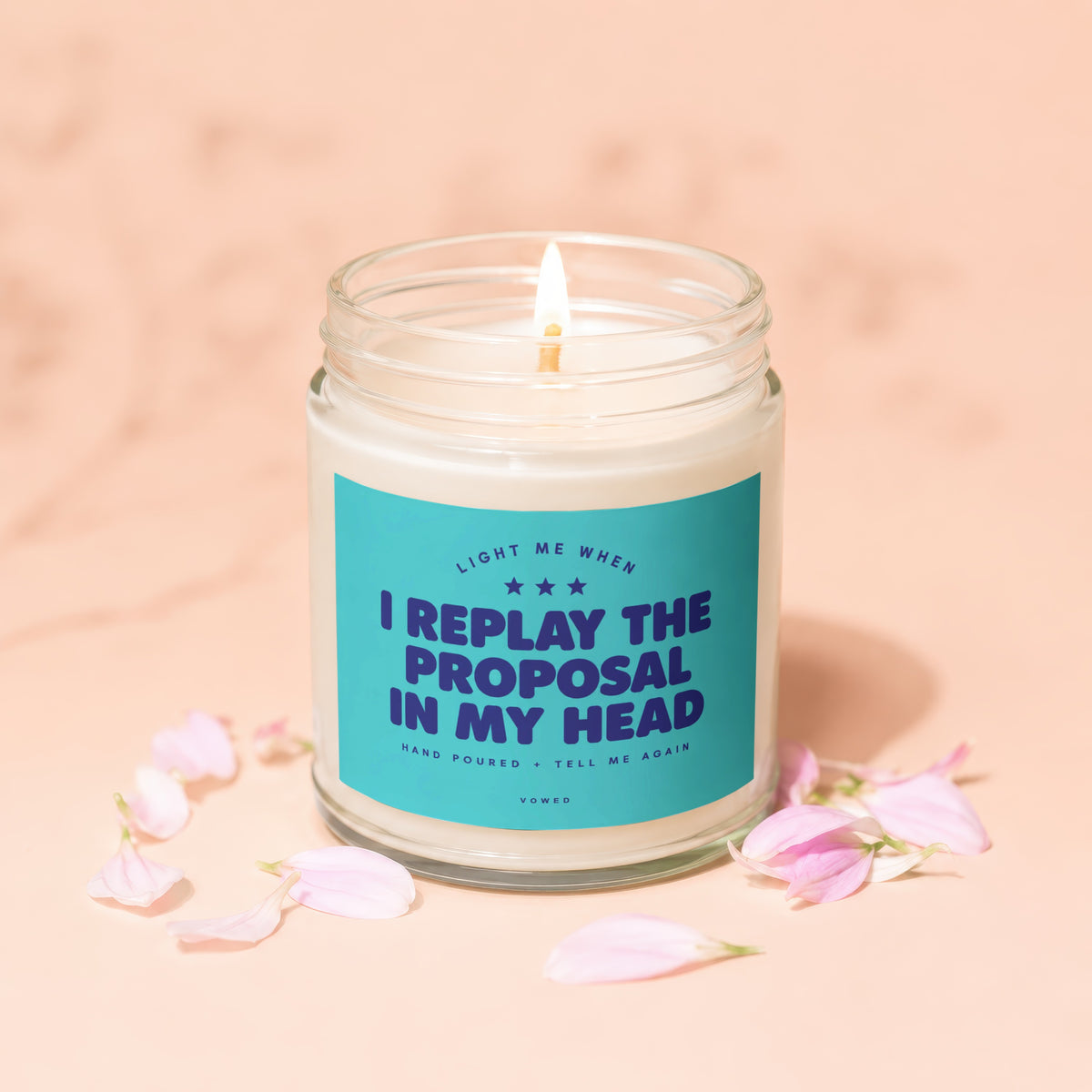 Lit candle with "I REPLAY THE PROPOSAL IN MY HEAD" on label, surrounded by petals.