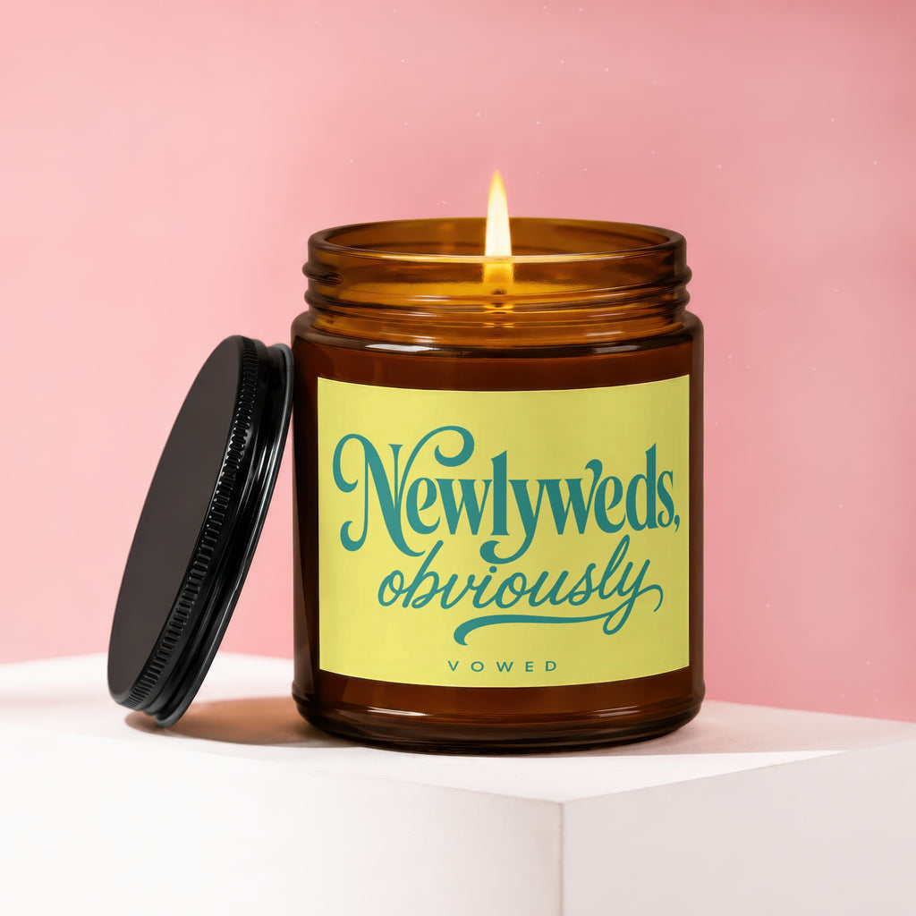 Amber jar candle with lit wick and black lid, features "Newlyweds, obviously" text.