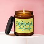 Amber jar candle with lit wick and black lid, features "Newlyweds, obviously" text.