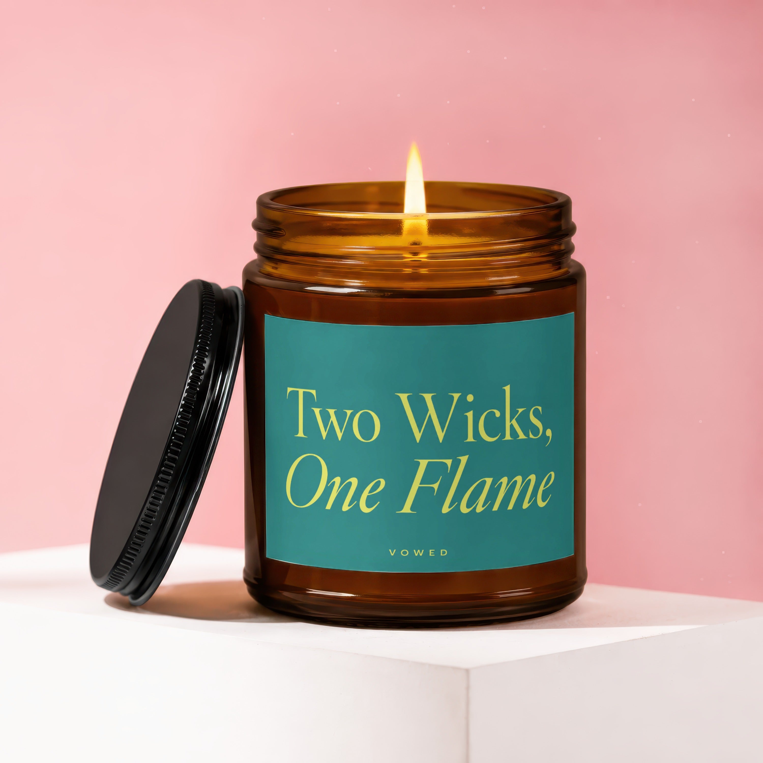 Amber jar candle with lit wick and teal label reading "Two Wicks, One Flame"
