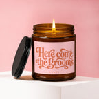 Lit amber jar candle with pink label reading "Here come the grooms"