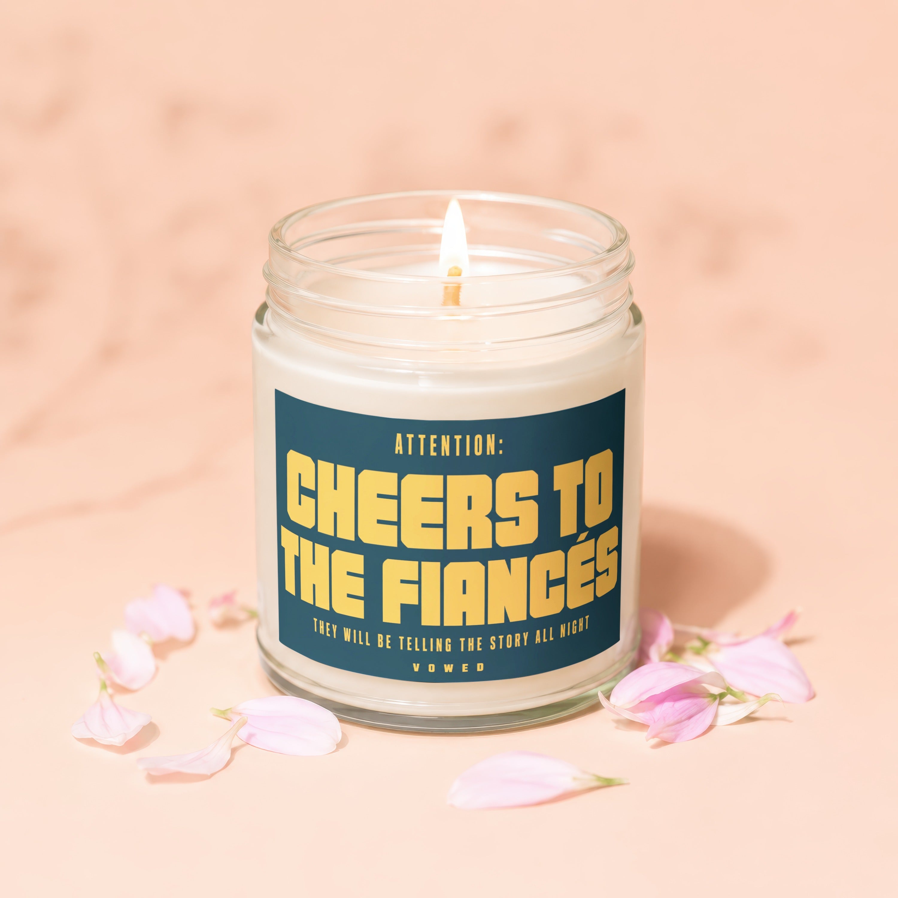 Candle with gold text: "Attention: Cheers to the Fiances", pink petals scattered.