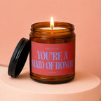Amber jar candle, flickering flame, with red label reading "Smells Like You're A Maid Of Honor".