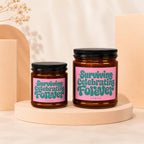 Two brown candles with black lids and pink labels that read "Surviving Celebrating Forever".