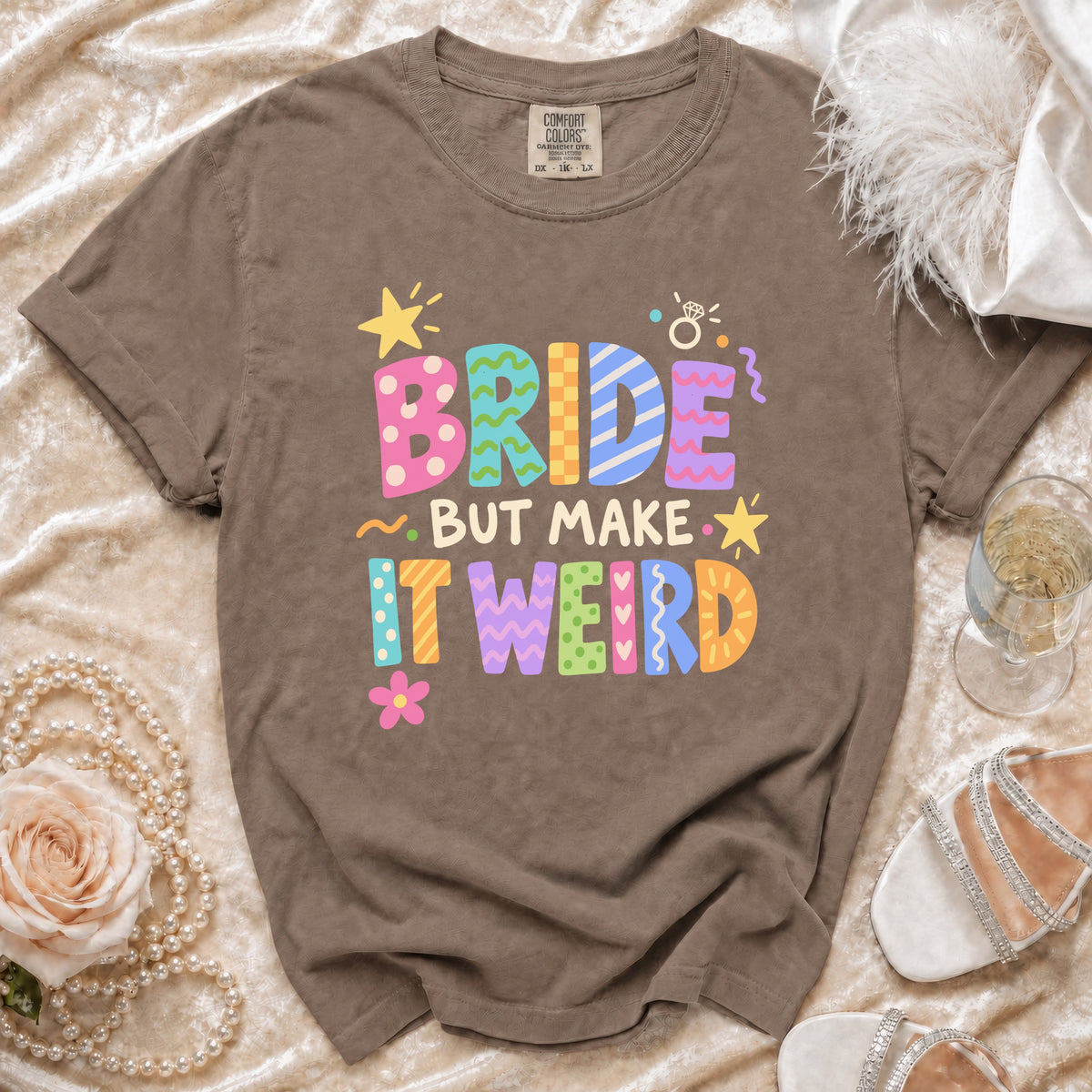 Brown t-shirt with colorful "BRIDE BUT MAKE IT WEIRD" text and bridal accessories.