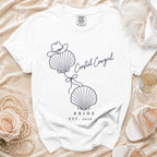 White t-shirt with a blue graphic of two seashells tied with a bow, a cowboy hat, and text reading Coastal Cowgirl Bride Est. 2026.
