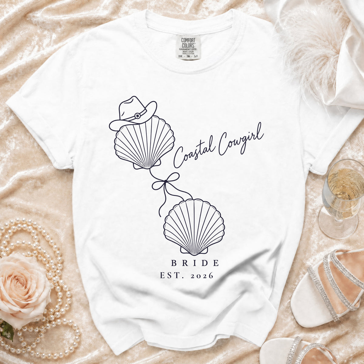 White t-shirt with a blue graphic of two seashells tied with a bow, a cowboy hat, and text reading Coastal Cowgirl Bride Est. 2026.