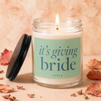 Lit candle in glass jar with "it's giving bride" text, lid off to the side.