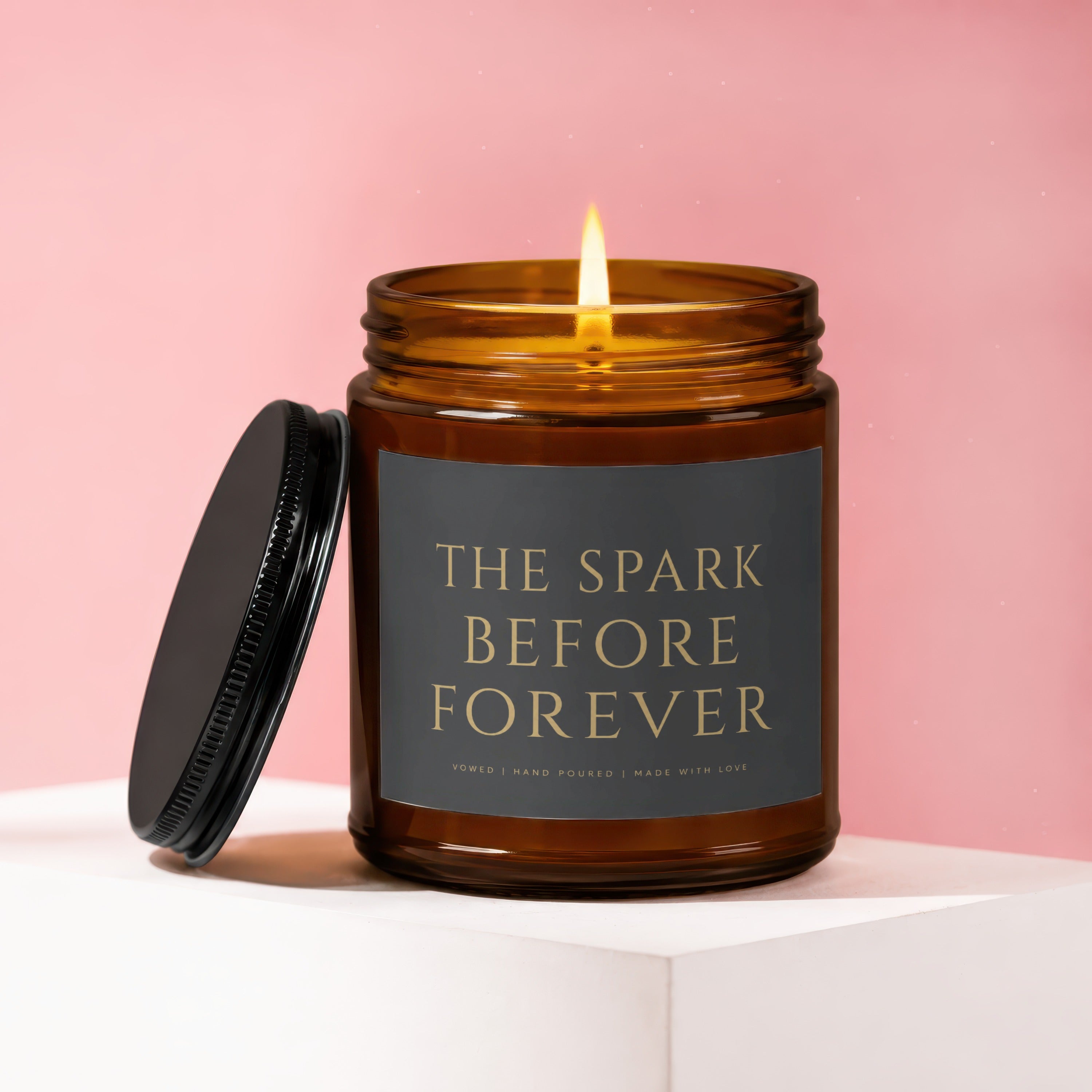 Amber candle with lit wick, black lid beside it. Label reads "The Spark Before Forever."