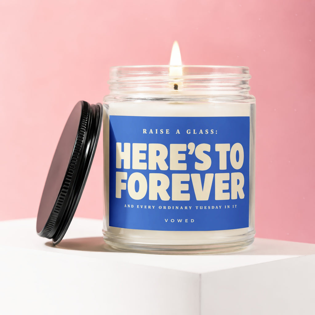 Scented candle in jar with black lid, reads "HERE'S TO FOREVER".