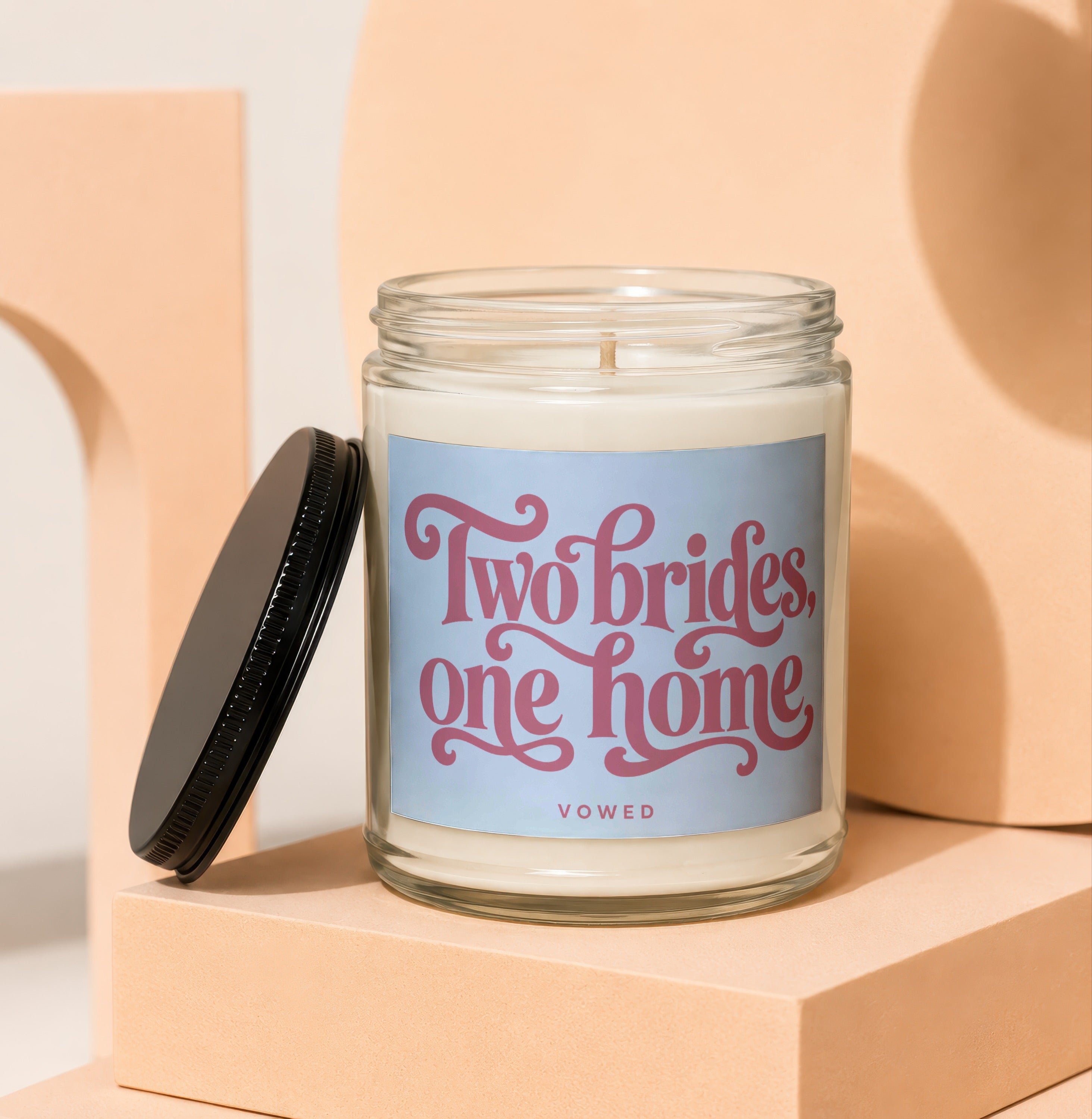 Glass candle with "Two brides, one home" and "vowed" in pink text on blue label.