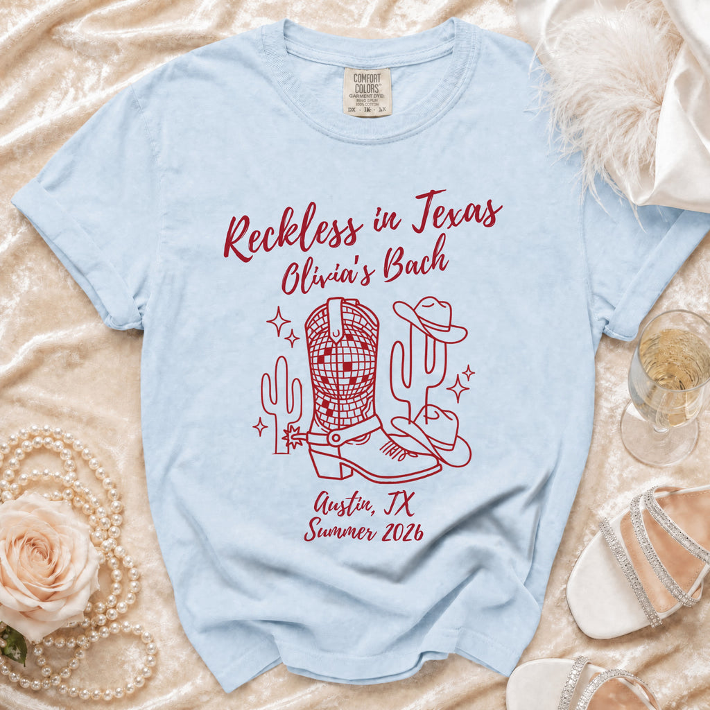 Light blue t-shirt with red text and graphics: "Reckless in Texas", cowboy hat, cactus, boots, and "Olivia's Bach".