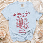 Light blue t-shirt with red text and graphics: "Reckless in Texas", cowboy hat, cactus, boots, and "Olivia's Bach".