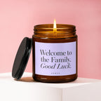 Amber candle with lit wick, lid off, labeled "Welcome to the Family. Good Luck. VOWED."