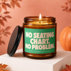 Amber jar candle with lit flame, green label reads "NO SEATING CHART. NO PROBLEM."