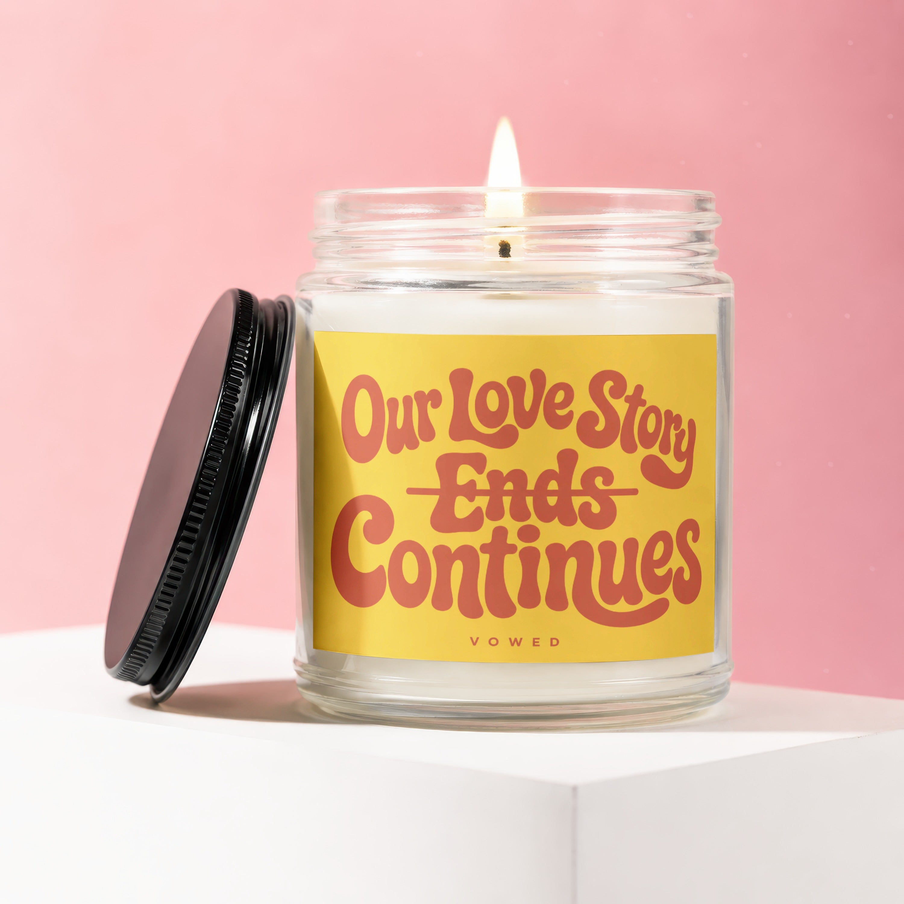 Glass candle with lit wick and yellow label reading "Our Love Story Ends Continues".