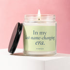 Candle in glass jar with lit wick and black lid off to the side with text "In my last-name-changing era."