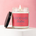 Glass jar candle with a lit wick and a pink label that reads "Good Luck, Babe. Vowed".