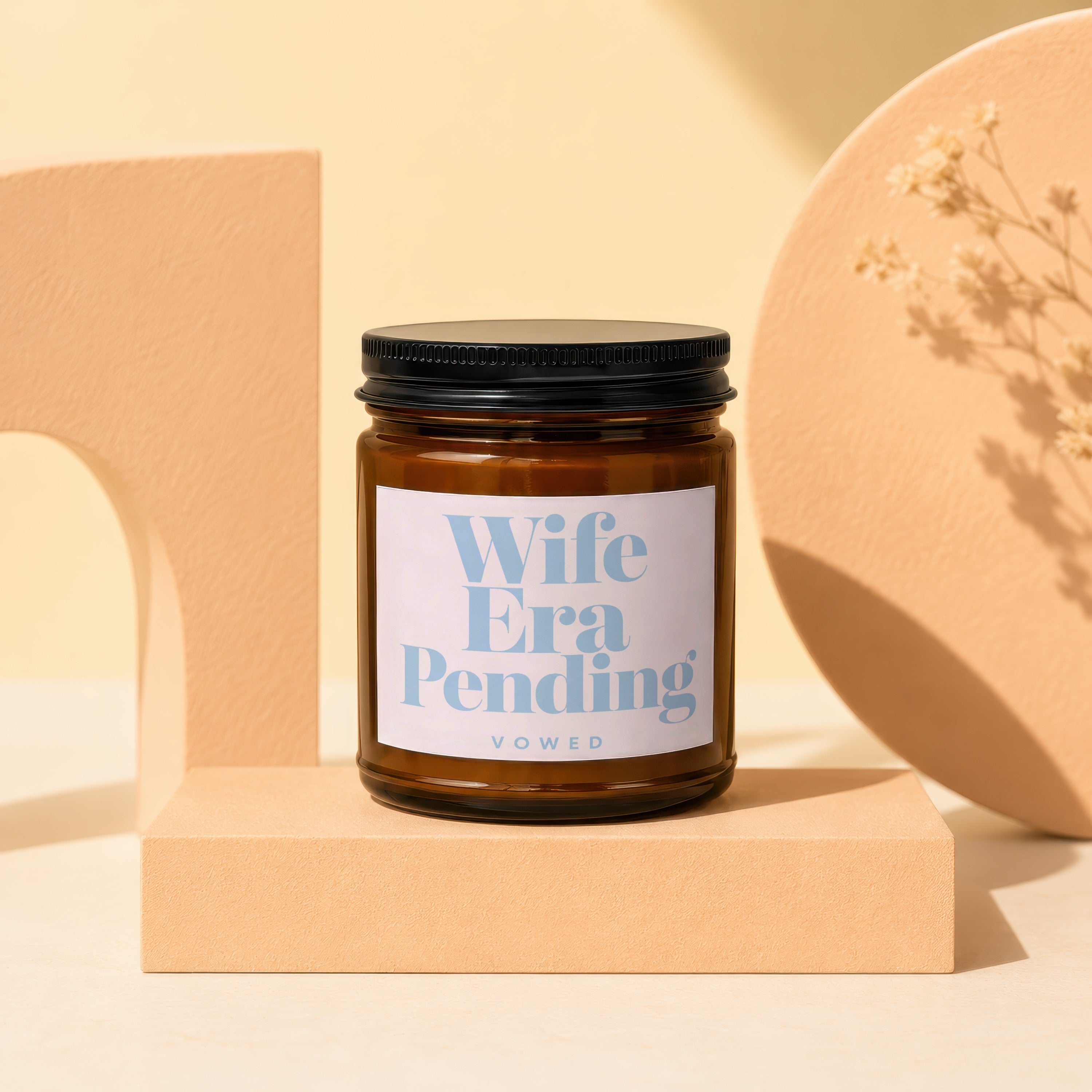 Amber jar candle with "Wife Era Pending VOWED" label resting on a cube.
