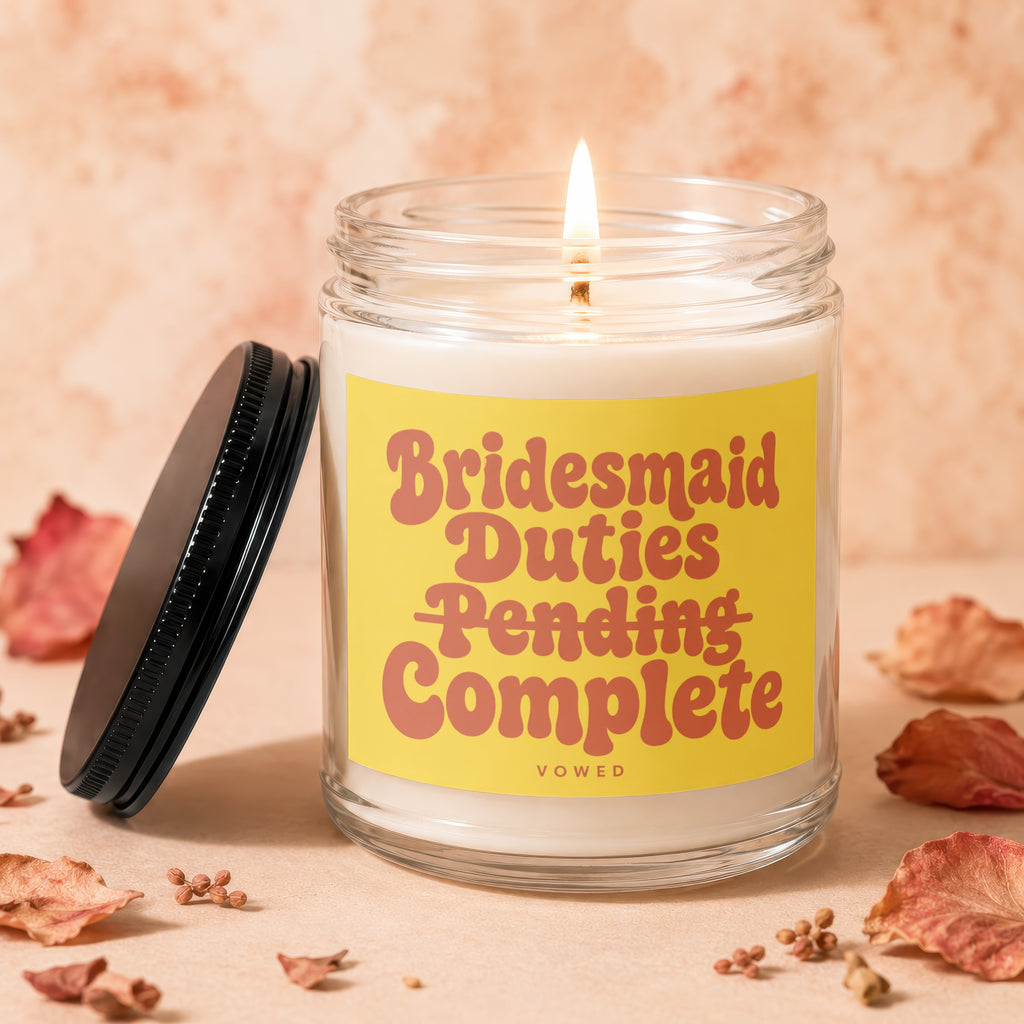 Glass candle with lit wick and yellow label reading "Bridesmaid Duties Pending Complete".