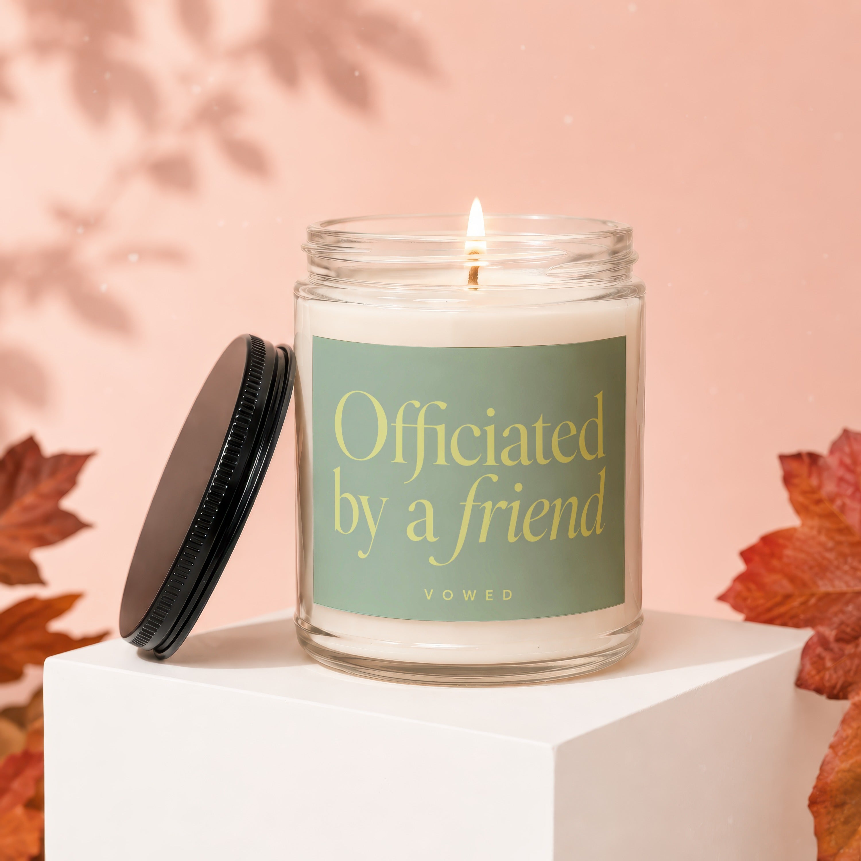 Glass candle with a lit wick and a green label reading "Officiated by a friend"