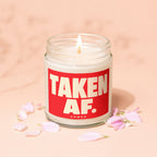 Jar candle with a lit wick and "TAKEN AF." text on a red label, surrounded by petals.