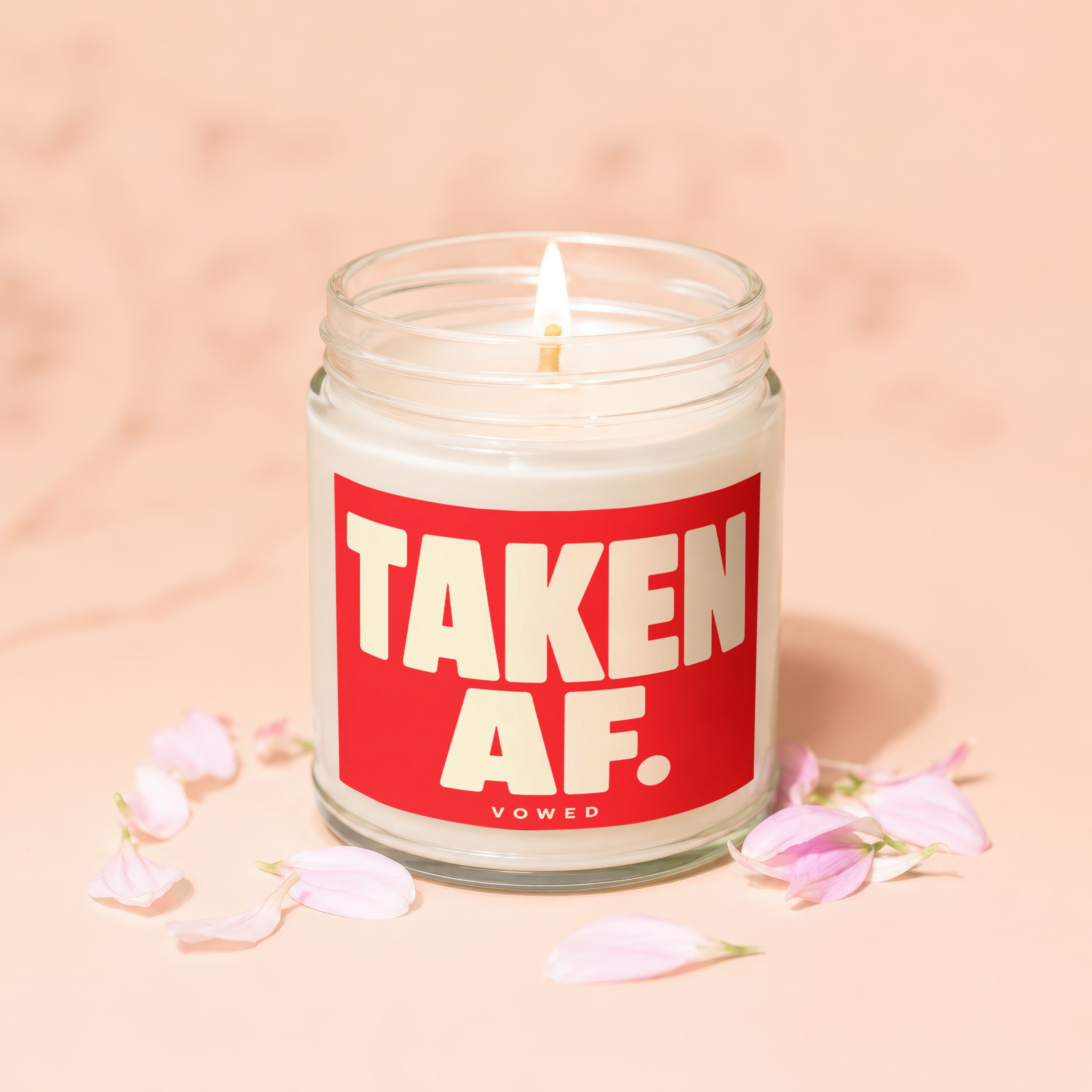 Jar candle with a lit wick and "TAKEN AF." text on a red label, surrounded by petals.